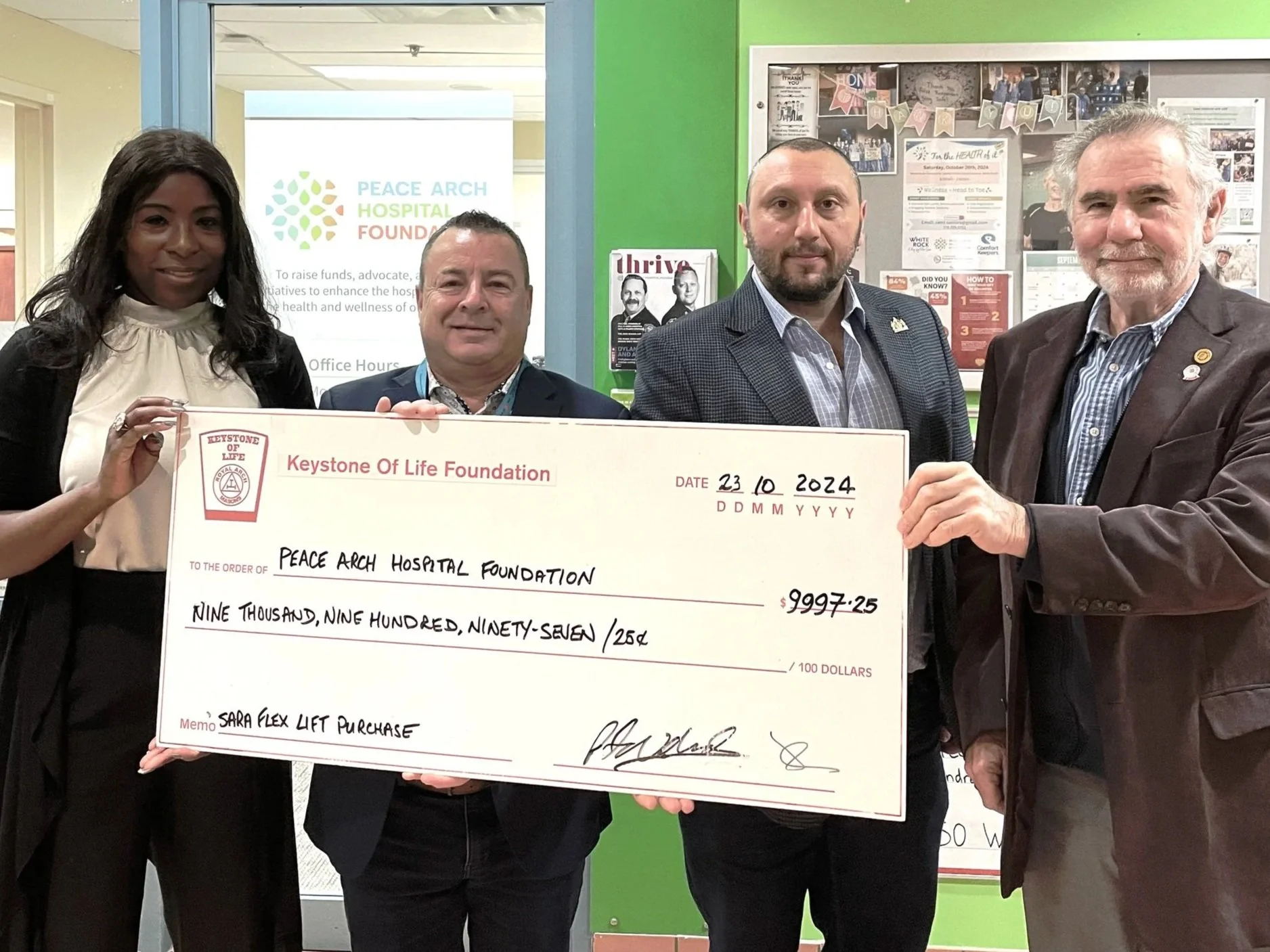 Four people are standing together indoors, holding a large ceremonial check made out to Peace Arch Hospital Foundation for $9,997.25. The group includes two men and two women, dressed in business casual attire. Behind them are bulletin boards with various notices and photos, and a sign that reads 'PEACE ARCH HOSPITAL FOUNDATION.'