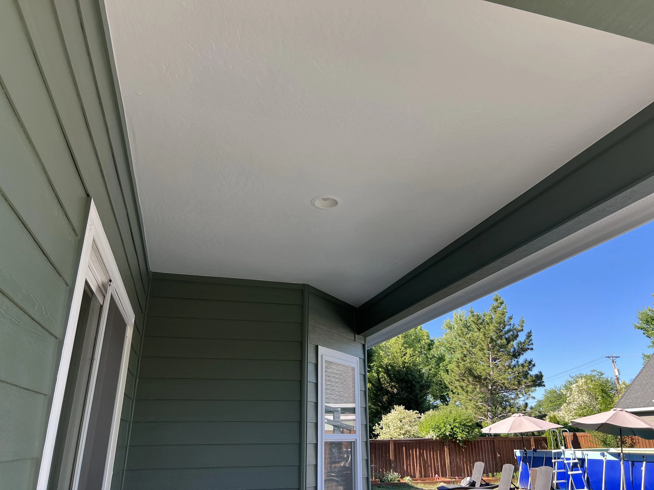 The image depicts the covered backyard of a house with green siding and a white ceiling. There are trees and outdoor furniture, including umbrellas and chairs, visible in the background under clear, sunny weather.