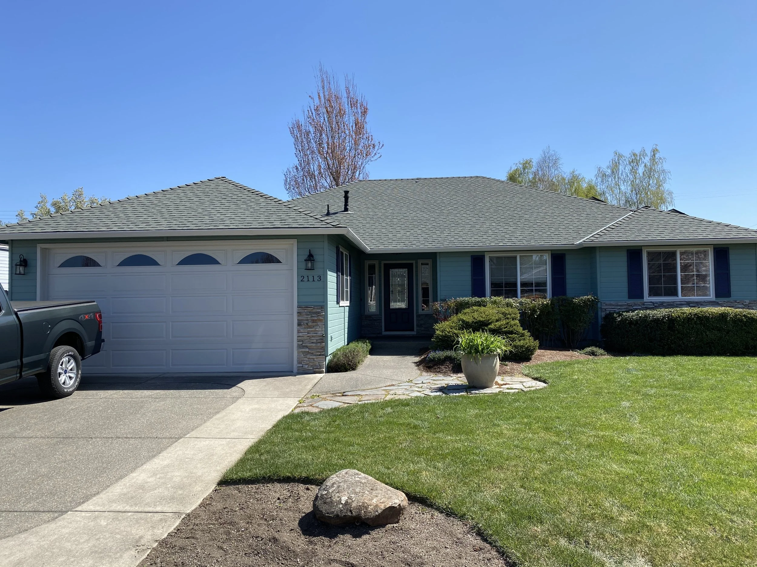 A suburban house with a light blue exterior, a white garage door, and a gray shingled roof. Front yard includes a curved concrete walkway, a large rock, a small garden bed, and a well-maintained lawn. A pickup truck is parked on the driveway, and tre