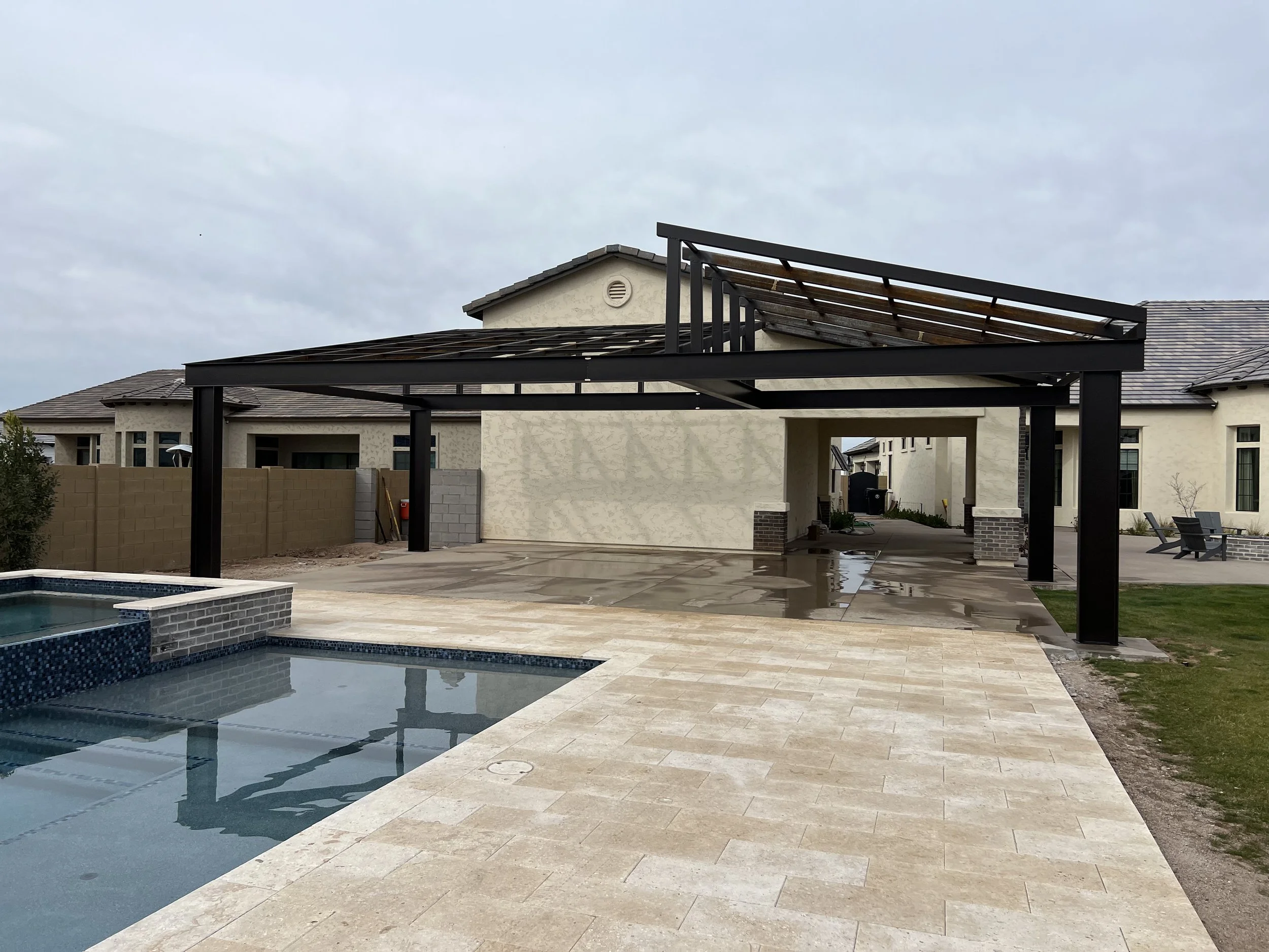 A backyard with a swimming pool, a beige house with a tiled roof, a covered patio area with a black frame, and cloudy sky overhead.