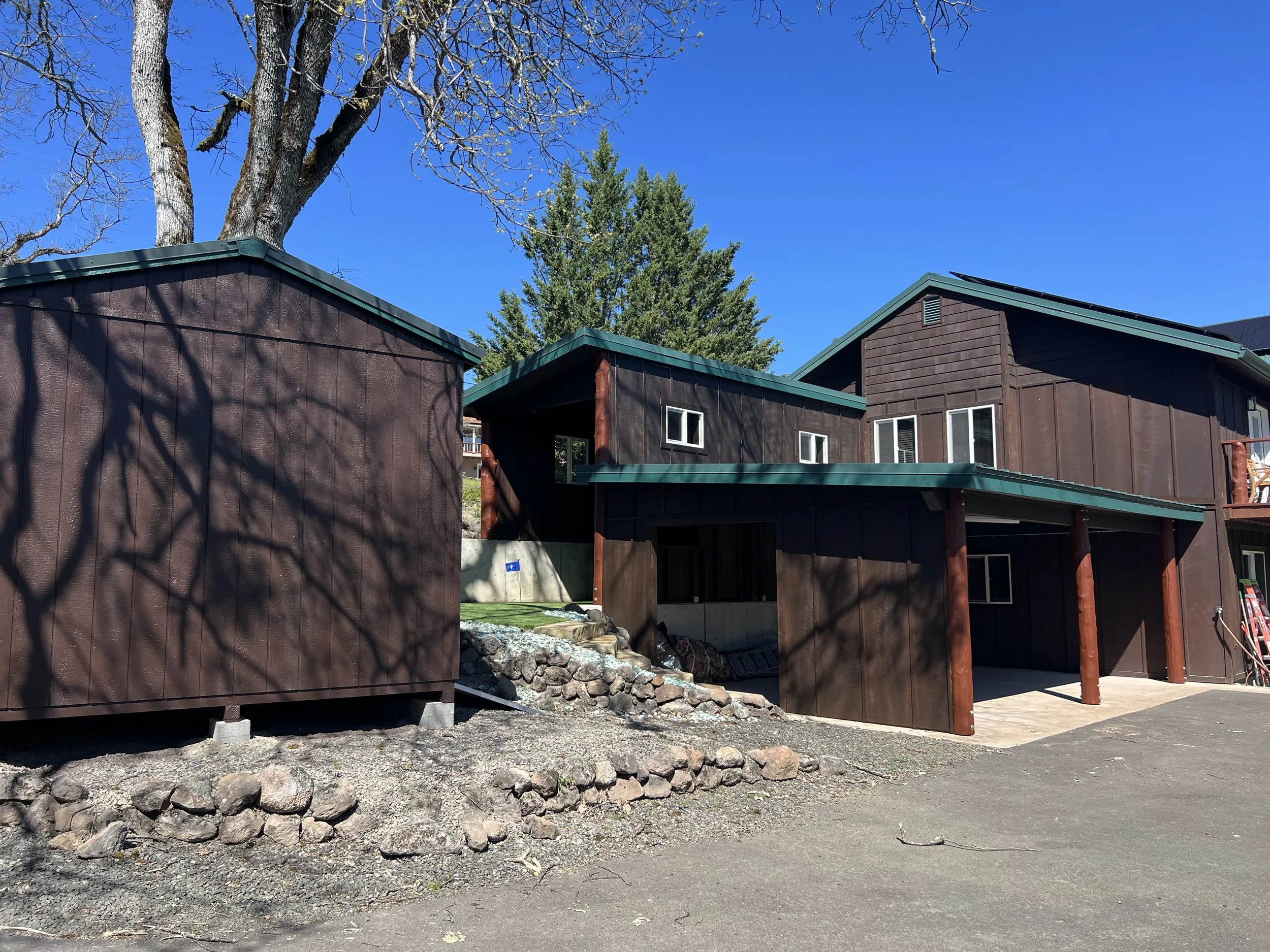 A residential house with dark brown siding and green roof, situated behind a stone and dirt yard with a large leafless tree and a tall evergreen tree in the background, under a clear blue sky.
