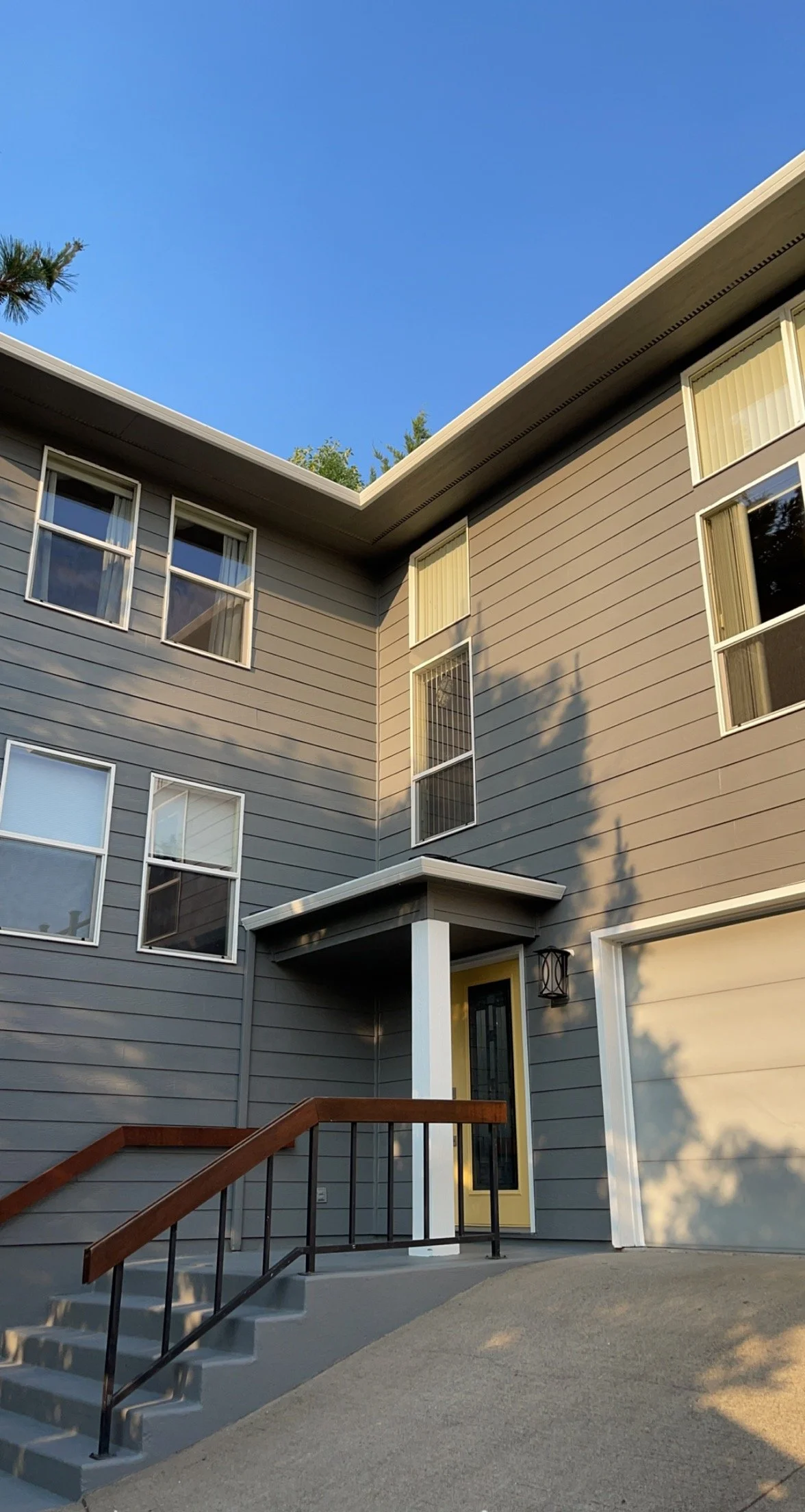 Front entrance of a multi-story residential building with gray siding, several windows, a small porch with stairs, and a wooden handrail. The sky is clear and blue with some trees casting shadows.