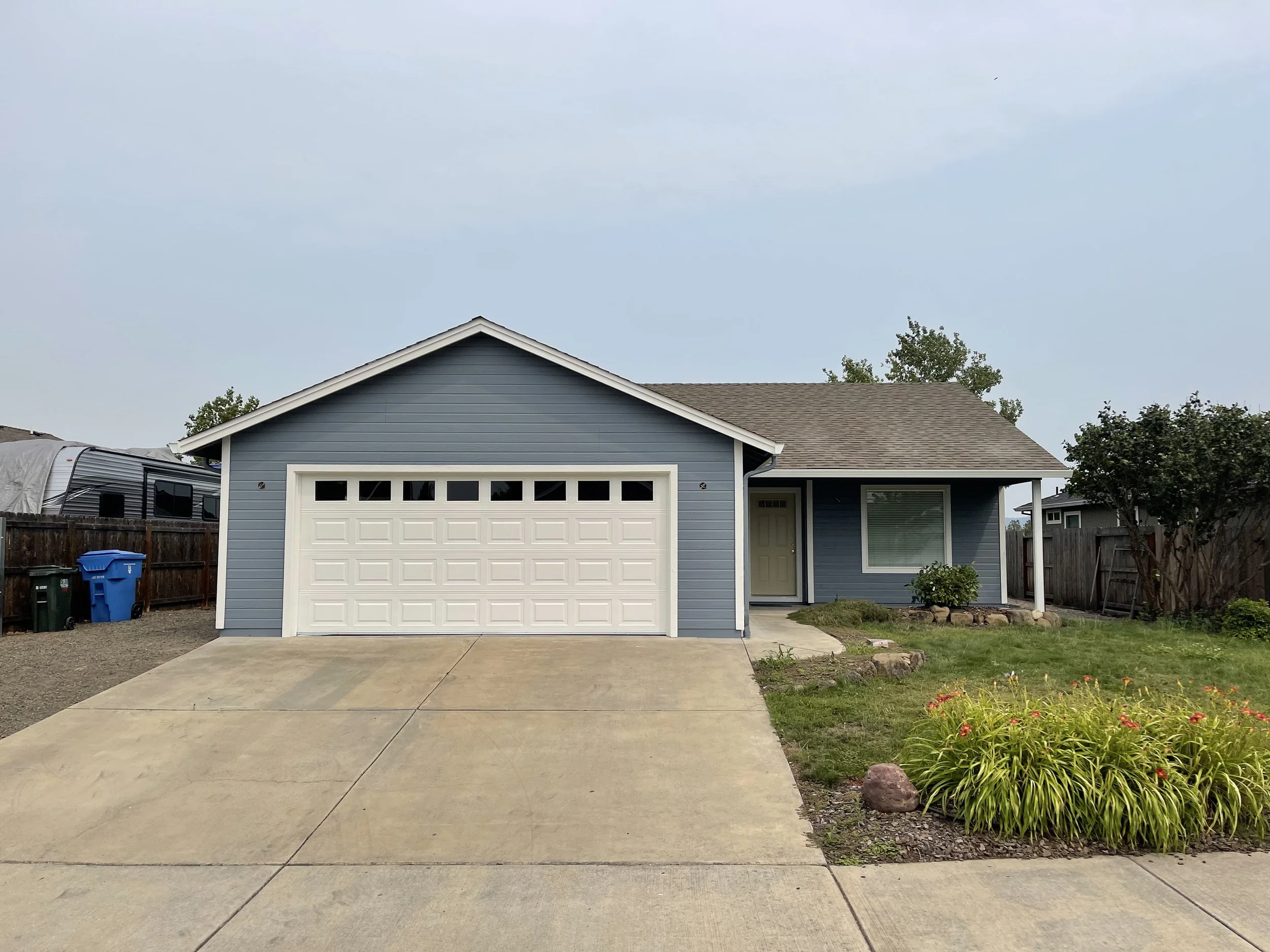 A blue house with a white garage door, front porch, and a green lawn with flowers. There are trees in the background and a cloudy sky.