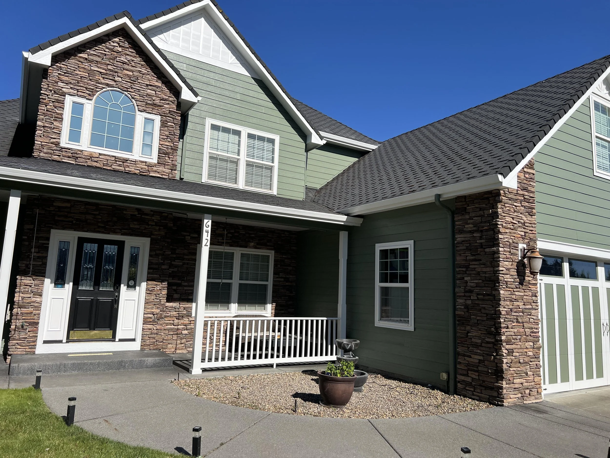 Front view of a two-story house with a gabled roof, green siding, and brick accents around the window and door. There is a small porch with a black front door, a white railing, and two planters with plants in front. The lawn is green, and the sky is 