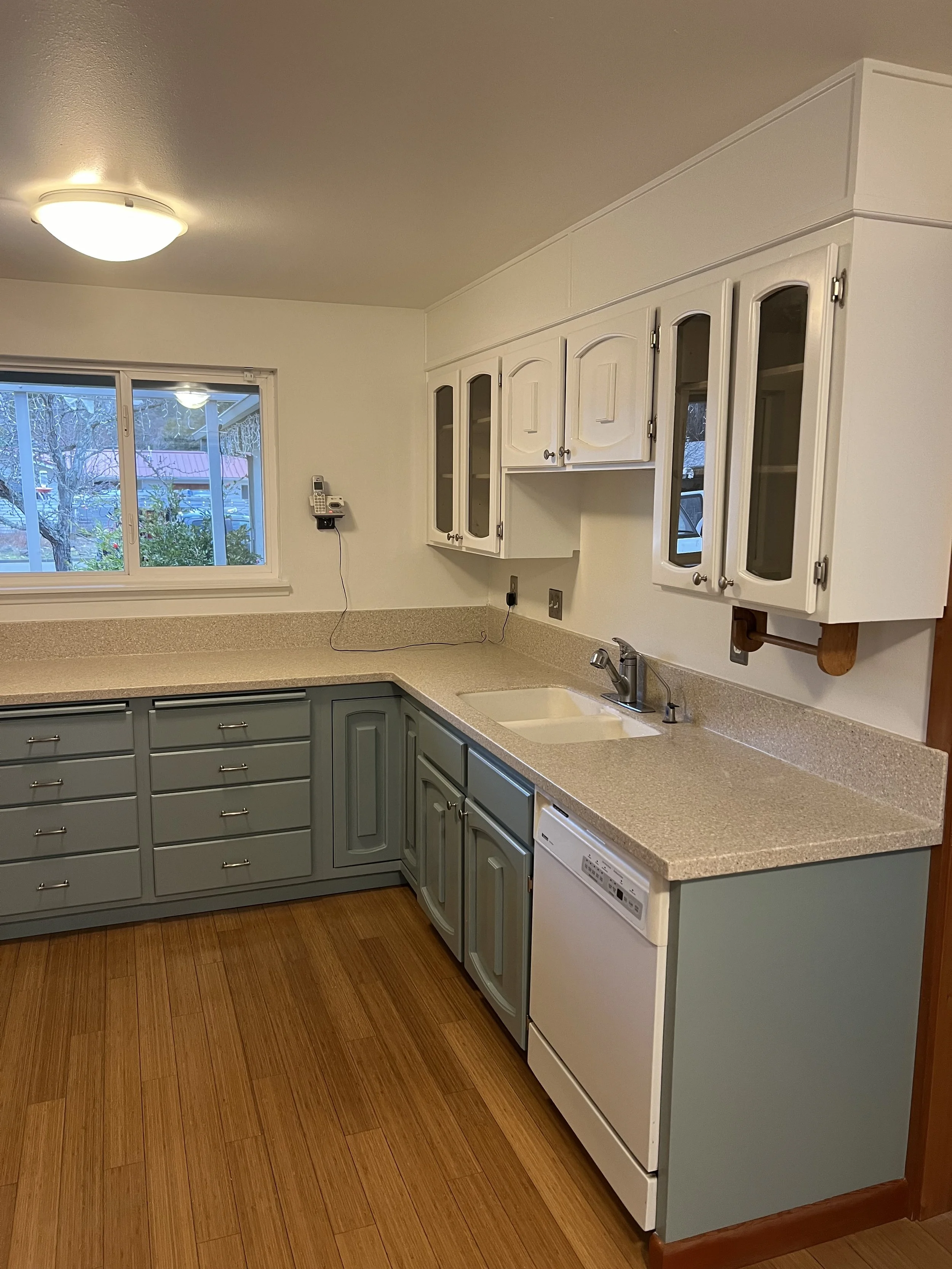 Kitchen with blue cabinets, beige countertops, white upper cabinets, window, and a backsplash with a light fixture on the ceiling.