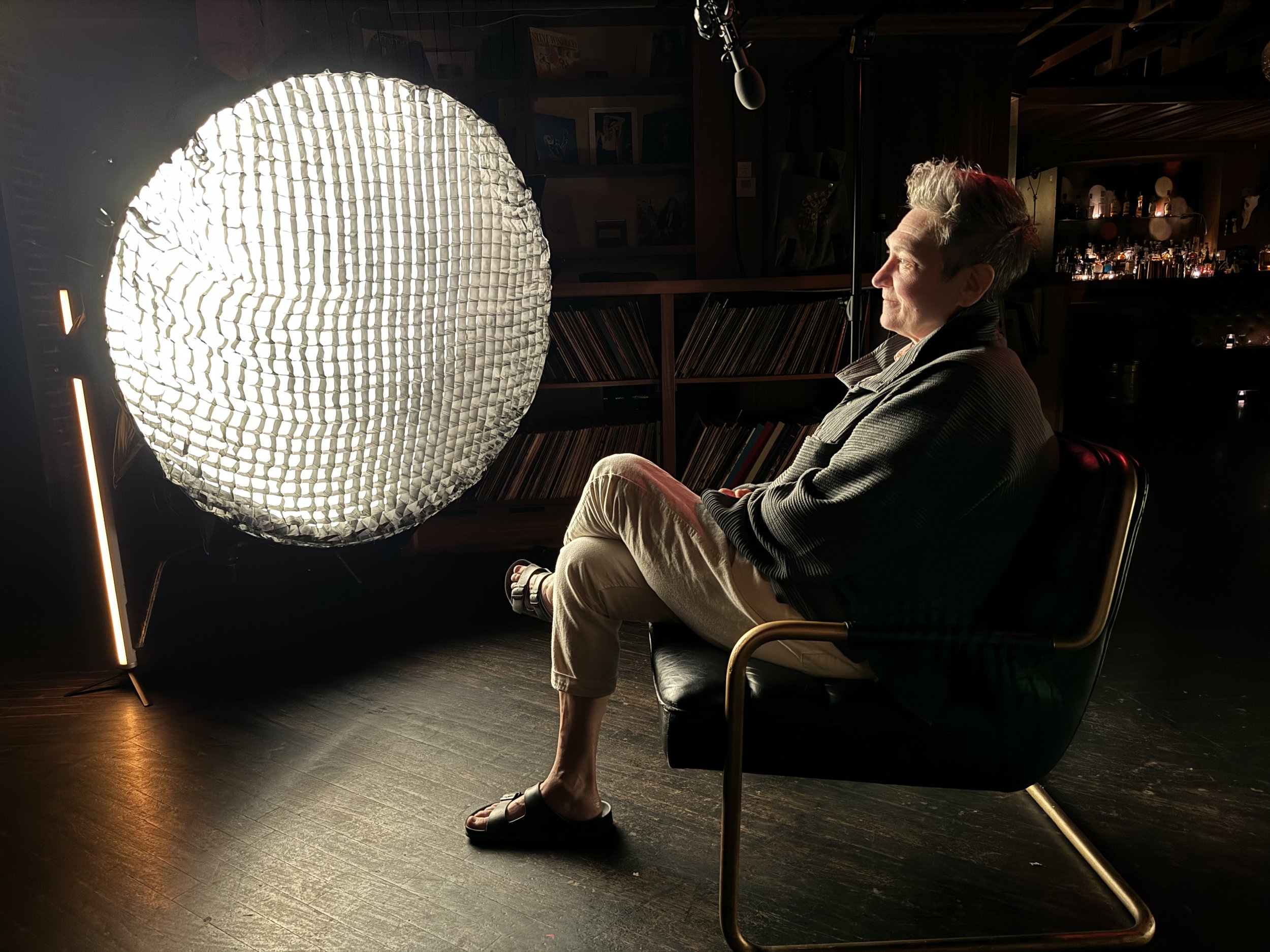 A woman sitting on a black chair in a dimly lit room, facing a bright, circular umbrella-style studio light. She is dressed in a striped jacket, beige pants, and sandals, with short gray hair. Shelves filled with vinyl records and bottles are visible in the background.