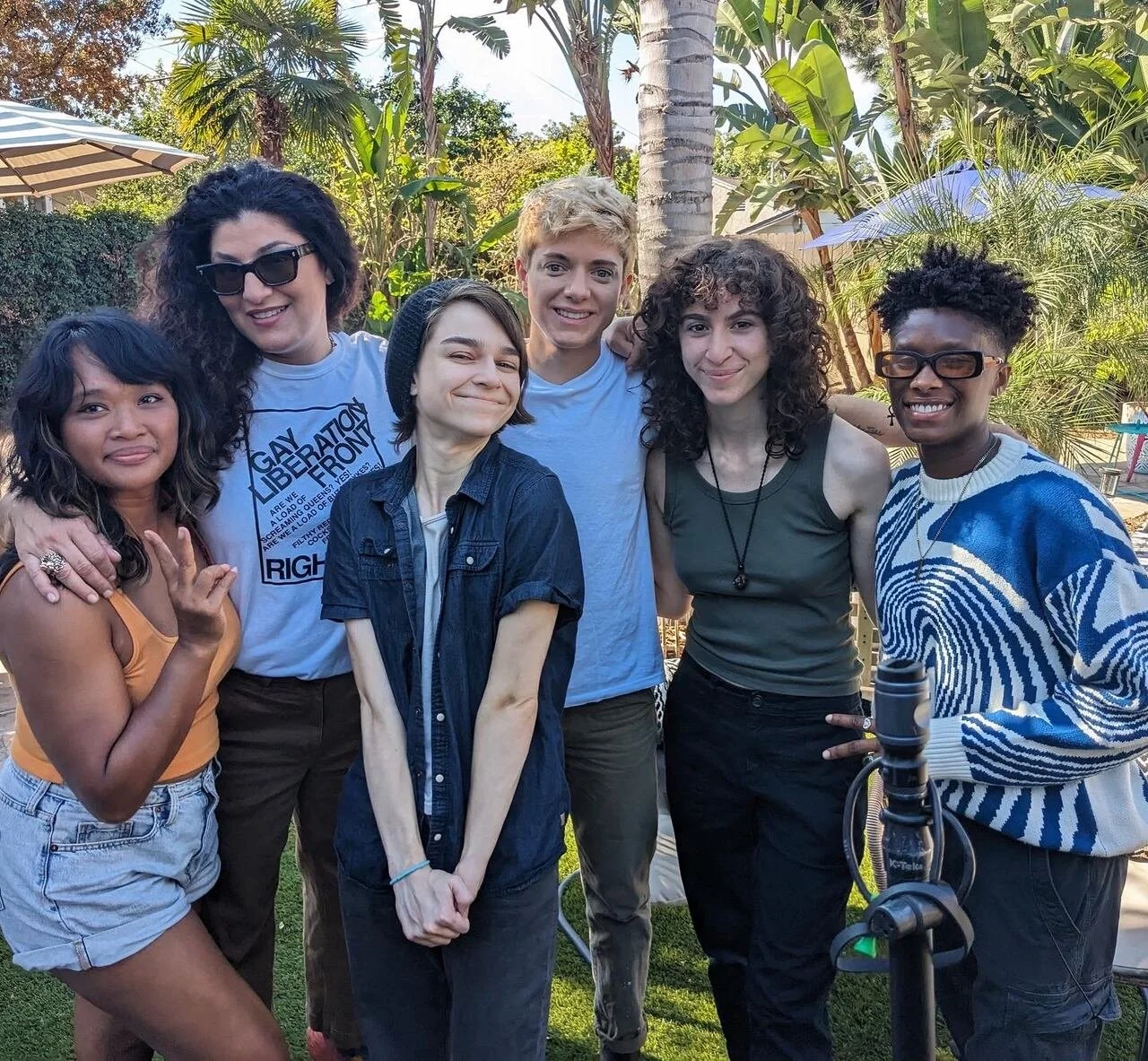 Group of six diverse people posing outdoors in a garden with trees and plants, smiling at the camera.