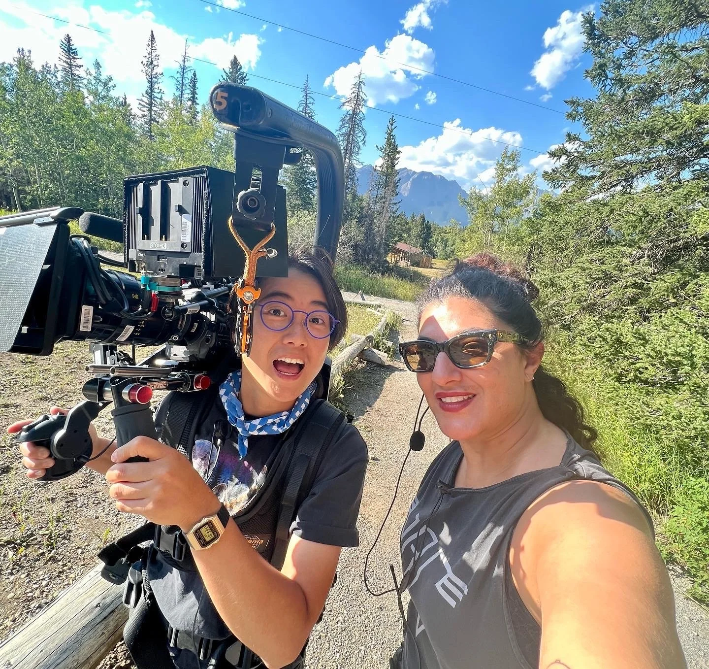Two people smiling outdoors with mountains, trees, and a blue sky behind them. One person is holding a professional camera and wearing glasses and a smartwatch. The other person is wearing sunglasses and a headset.