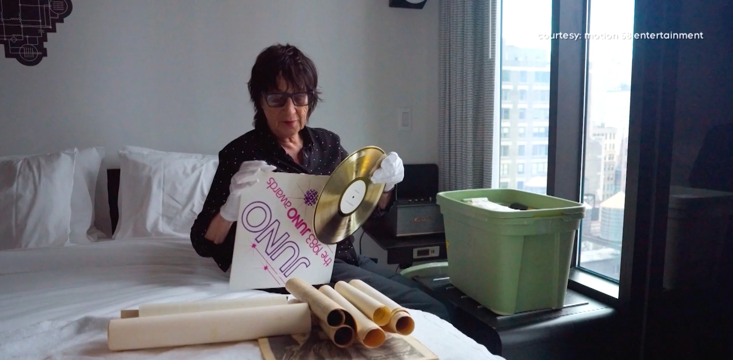 A woman sitting on a hotel bed holding a vinyl record and a cleaning instruction sign, with a green plastic bin and rolled-up papers nearby, in a room with city view through large window.