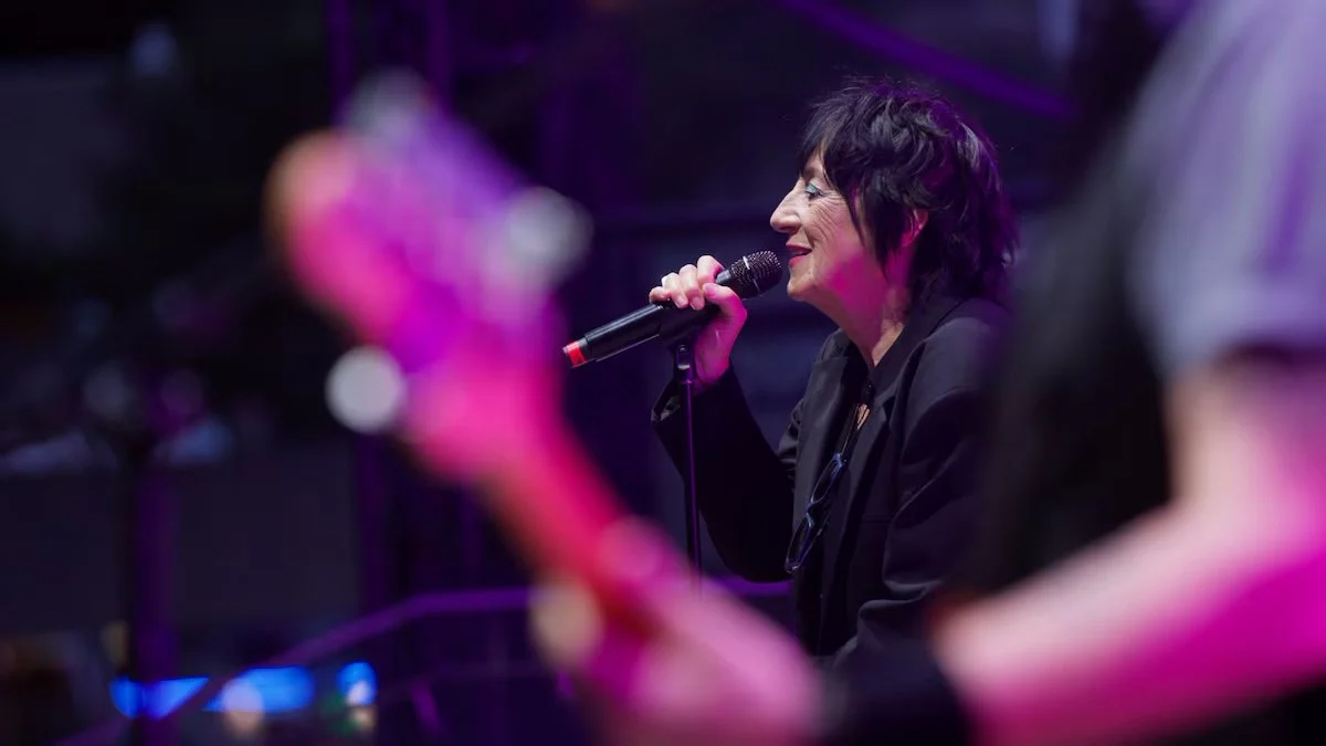 A woman with short black hair singing into a microphone on stage, with colorful stage lighting.