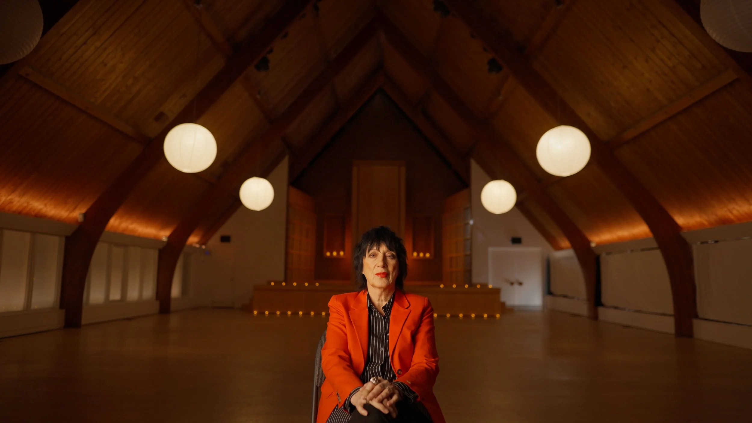 Woman with black hair and red lipstick sitting in a wooden room with vaulted ceiling and hanging lights.