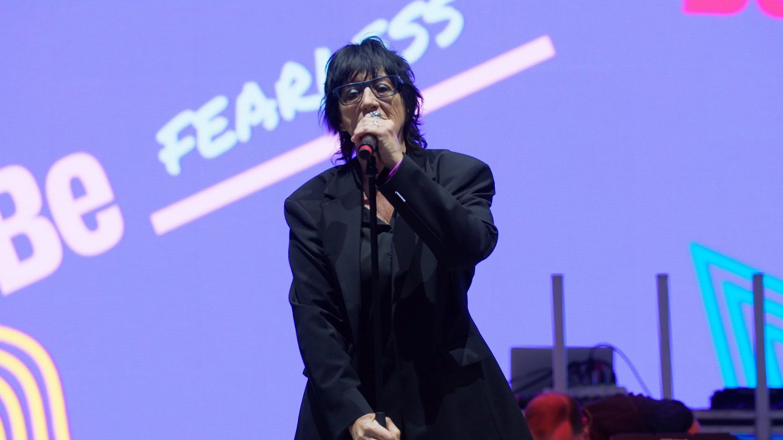 A woman with black hair, glasses, and wearing a black suit, holding a microphone, speaking on stage with a colorful digital background.