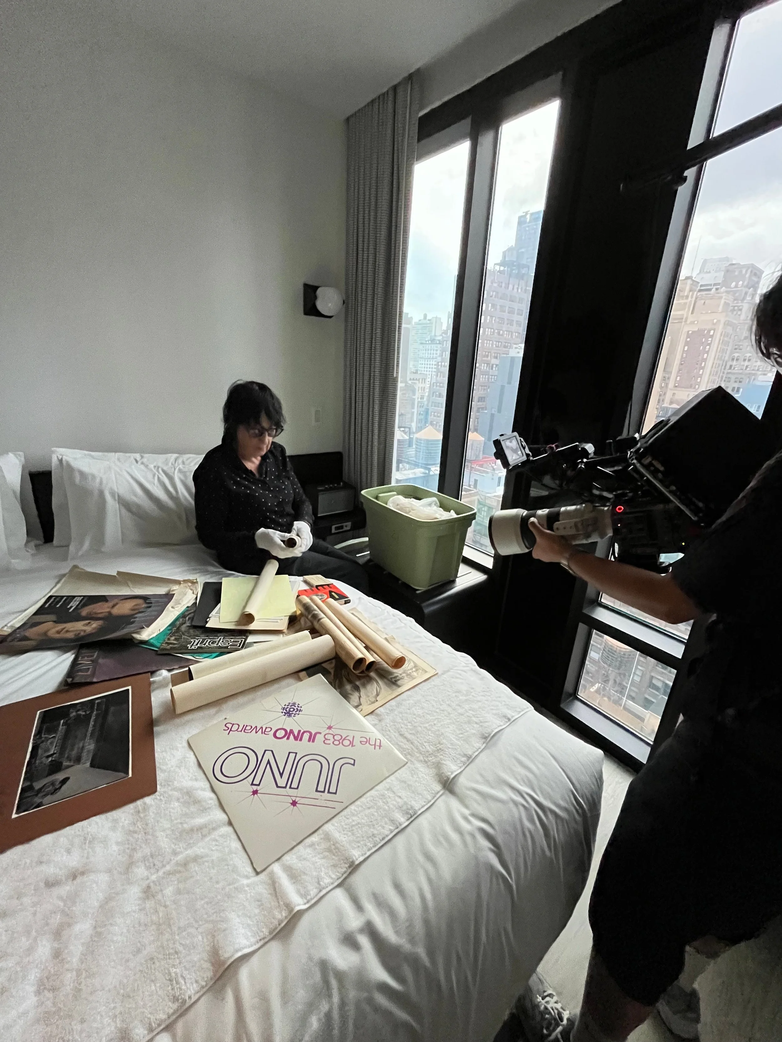 A woman sitting on a bed in a city apartment hotel room during daytime, holding a roll of paper, with art supplies, magazines, and posters spread on the bed, while a person with a camera records her, and tall buildings visible through large windows.