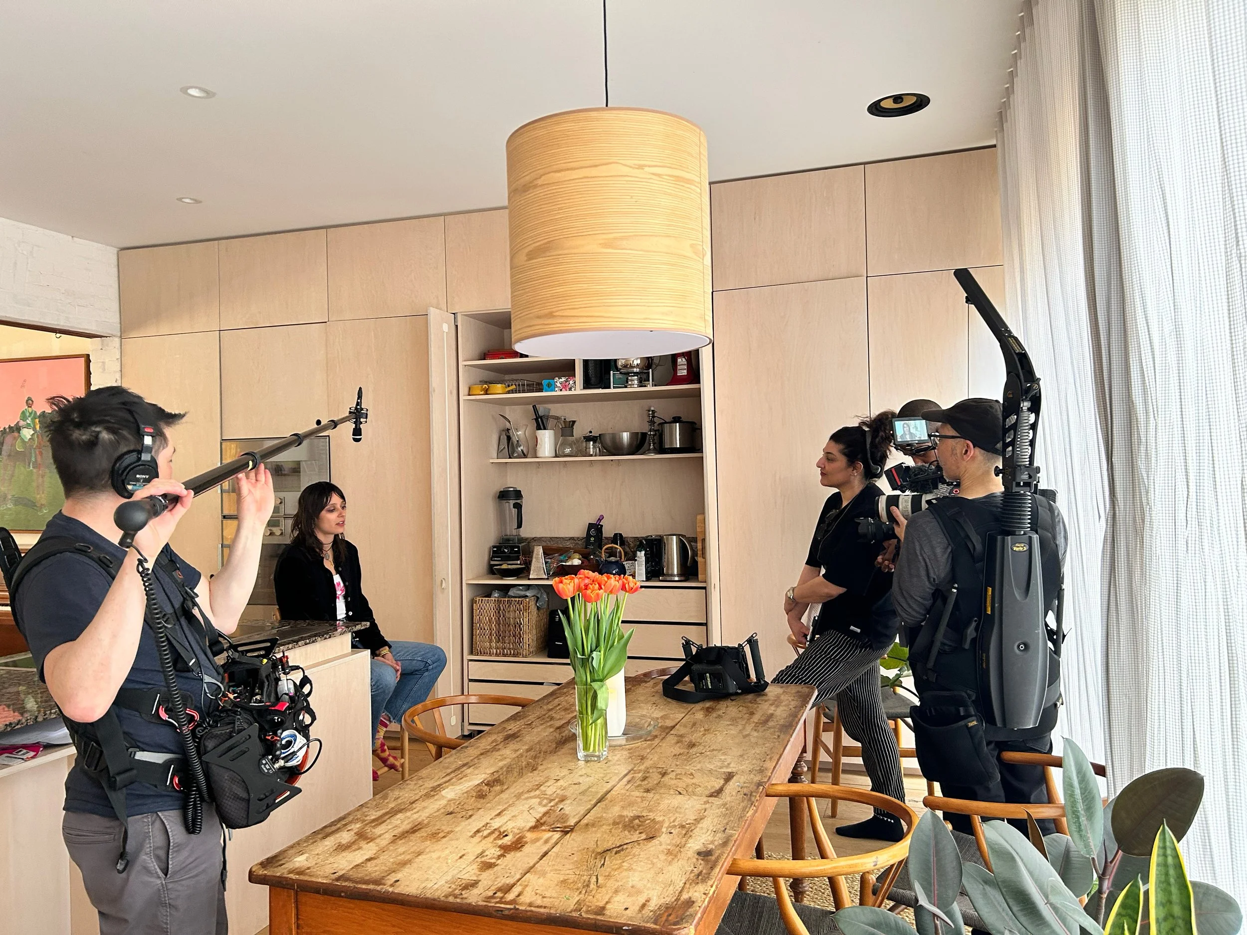 Filming crew setting up camera and microphone in a kitchen for a video interview with a woman sitting at a table with a vase of tulips.