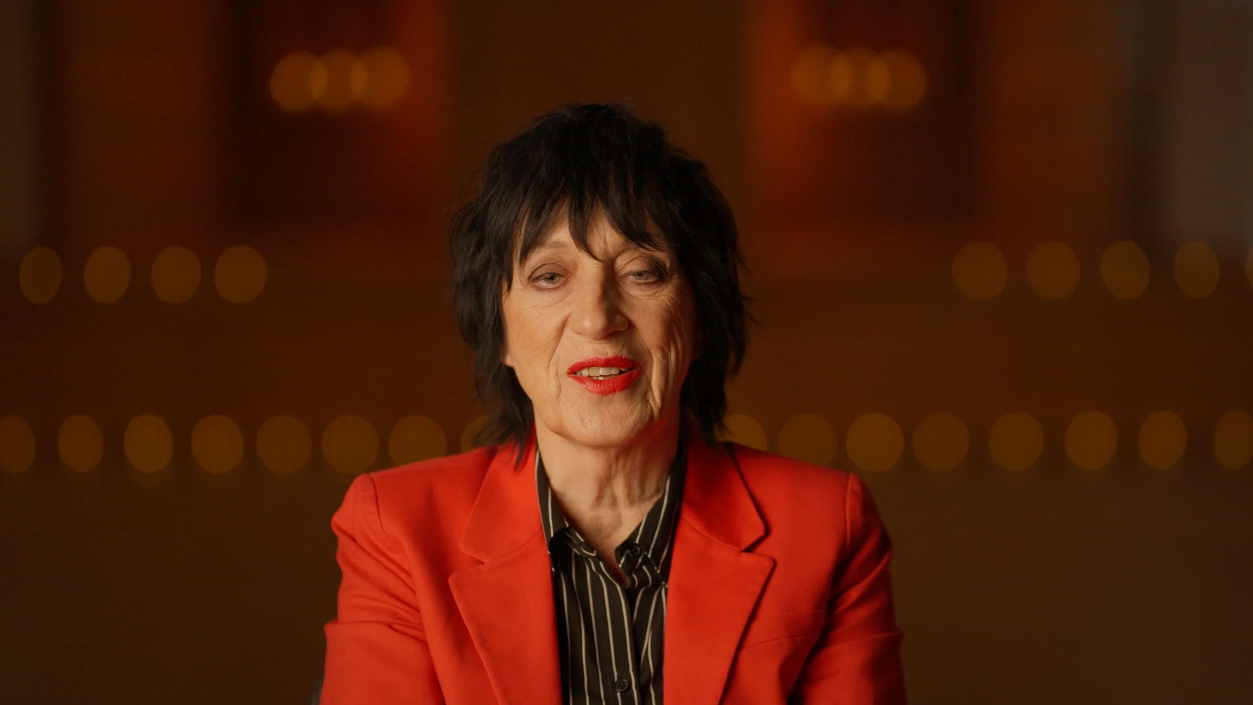 A woman with black, short, voluminous hair wearing a red blazer and black and white striped shirt, looking slightly to her left with a neutral expression against a background of blurred warm-colored lights.