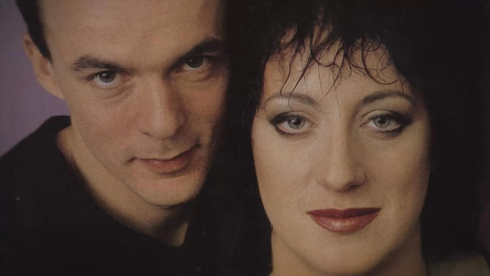 A close-up of a man and woman with intense expressions and dark clothing, looking directly at the camera.