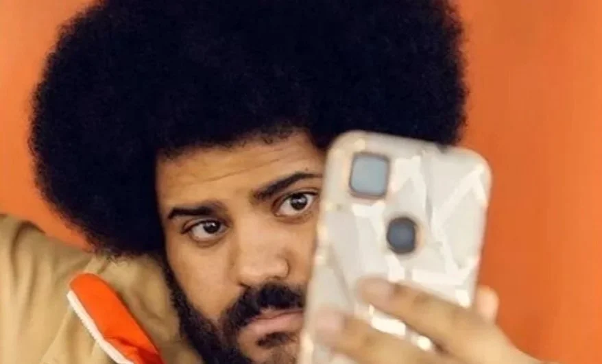 A man with an afro hairstyle and beard looking at his phone, with an orange background.