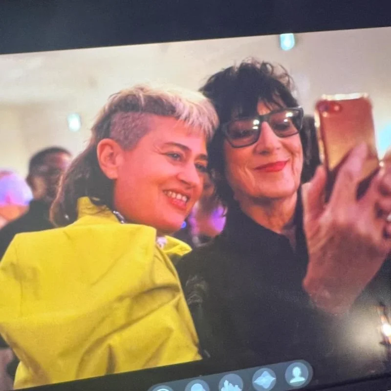 Two women taking a selfie at a social event. The woman on the left has short, blonde hair with darker roots and is wearing a yellow jacket. The woman on the right has dark, curly hair, wears glasses, and is dressed in black. There are other people in the background.