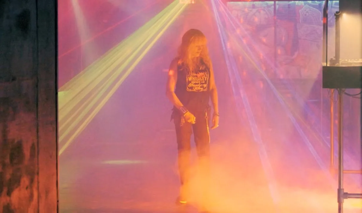 A woman with long hair stands in a colorful foggy nightclub with laser lights.