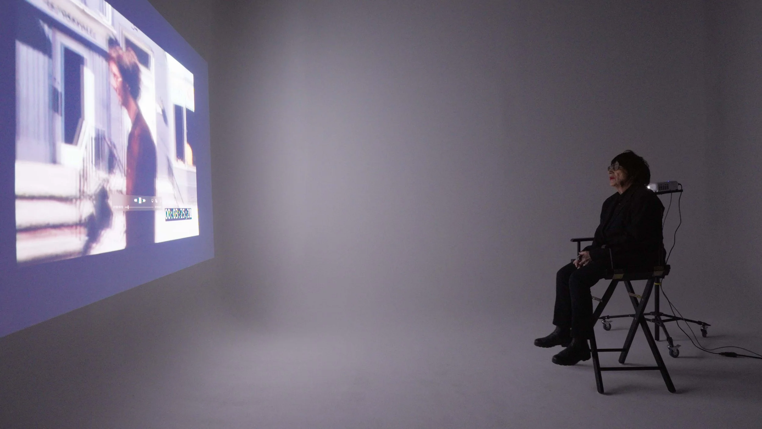 Woman sitting on chair observing a video projection on a wall, with a remote control in her hand, in a dimly lit room.