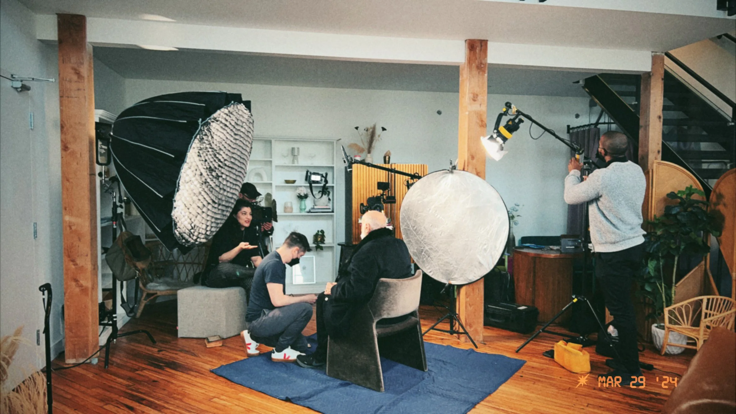 A film or photo shoot setup with lighting equipment, large reflectors, and crew members. Several people are sitting in front of cameras, engaging in conversation. The room has wooden floors, a blue rug, and a bookshelf with various decorative items.