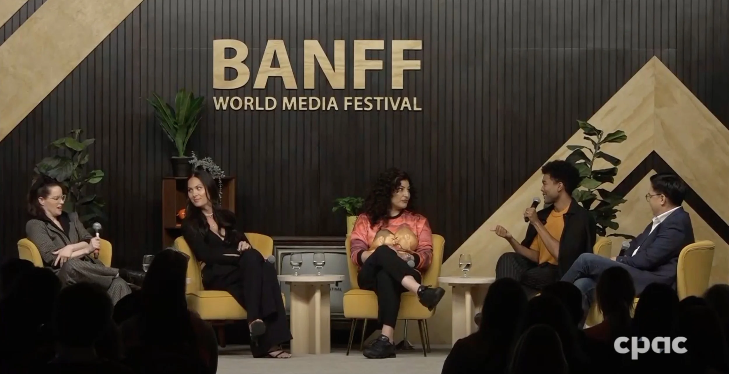 A panel discussion at Banff World Media Festival with five people sitting on yellow chairs on stage, engaging in conversation, with a dark textured background and large 'BANFF' sign.