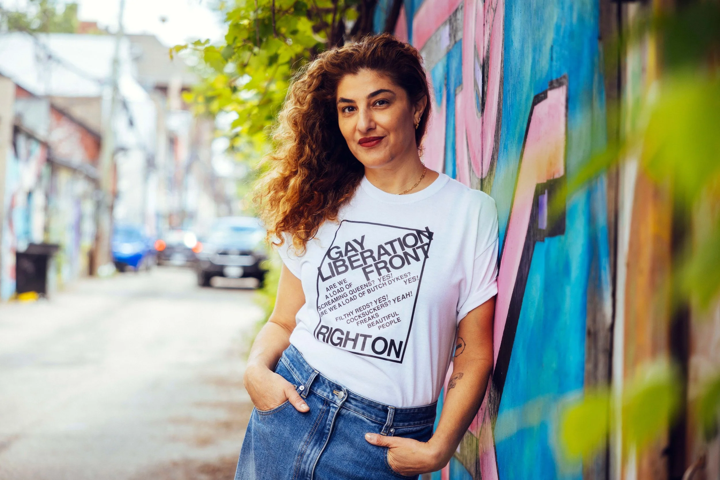A woman with curly hair wearing a white T-shirt with text, standing by a colorful mural on a city sidewalk with trees and parked cars in the background.