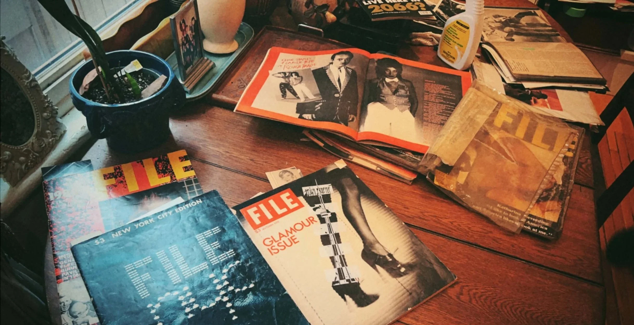 A cluttered wooden table holding magazines, newspapers, a potted plant, and miscellaneous items. There is a magazine titled 'FILE' and an open magazine with black-and-white photographs of people, along with a nightstand or windowsill with a small potted plant, a framed picture, and a tube of sunscreen.