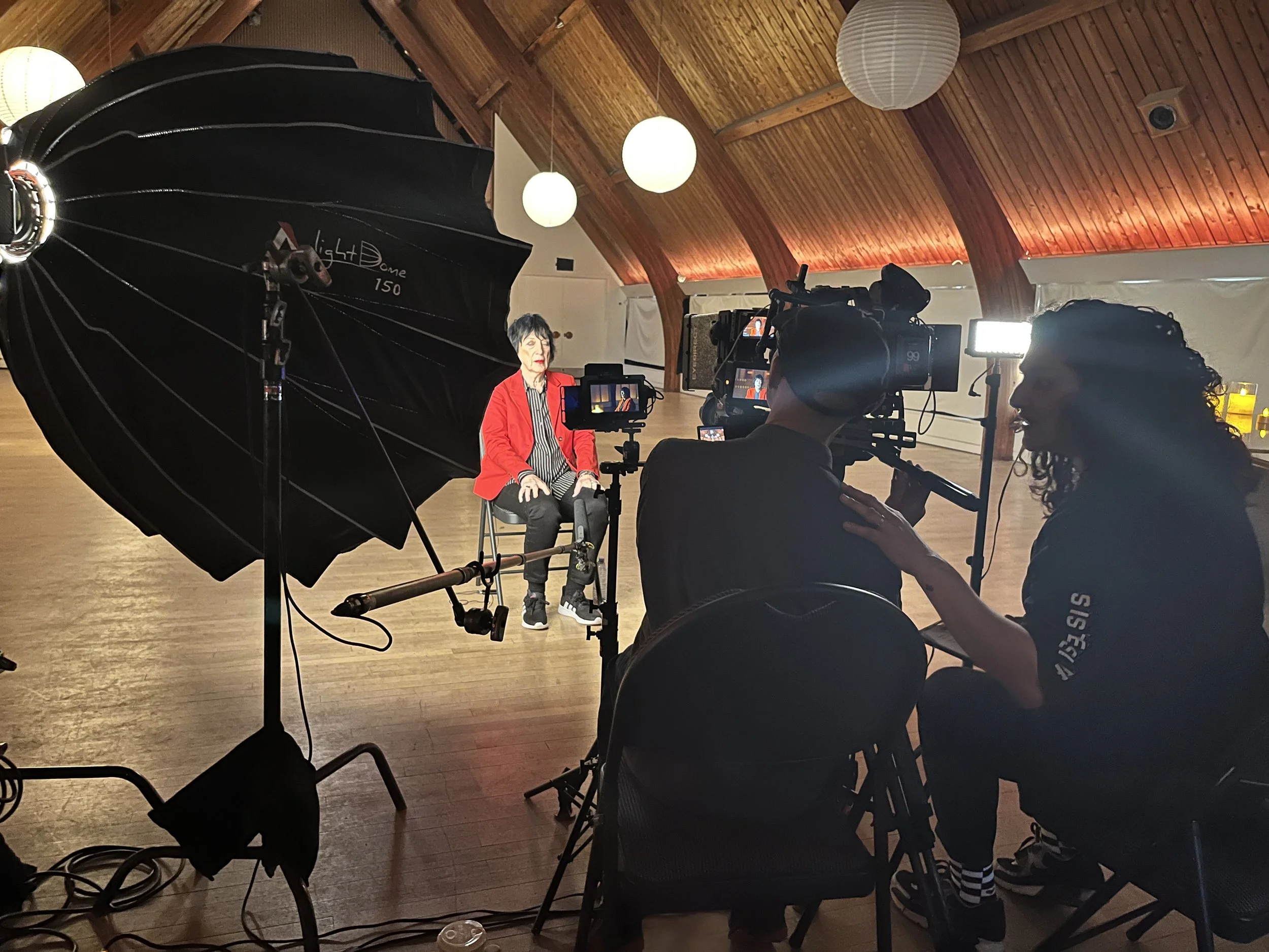 A woman in a red blazer and black pants being filmed with multiple cameras inside a wooden ceiling room with hanging paper lanterns.