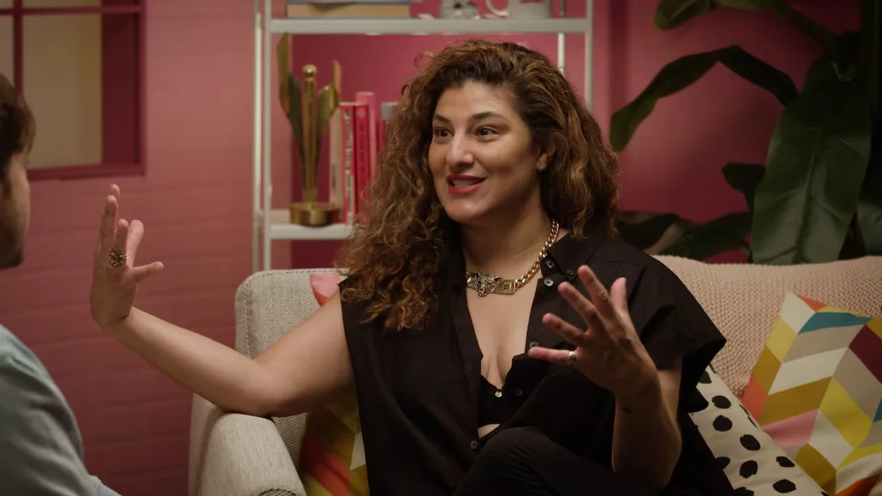 A woman with curly brown hair wearing a black shirt, gold jewelry, and sitting on a beige couch, engaging in conversation with a man. The background features pink walls, a white bookshelf with pink books, green plants, and colorful pillows.