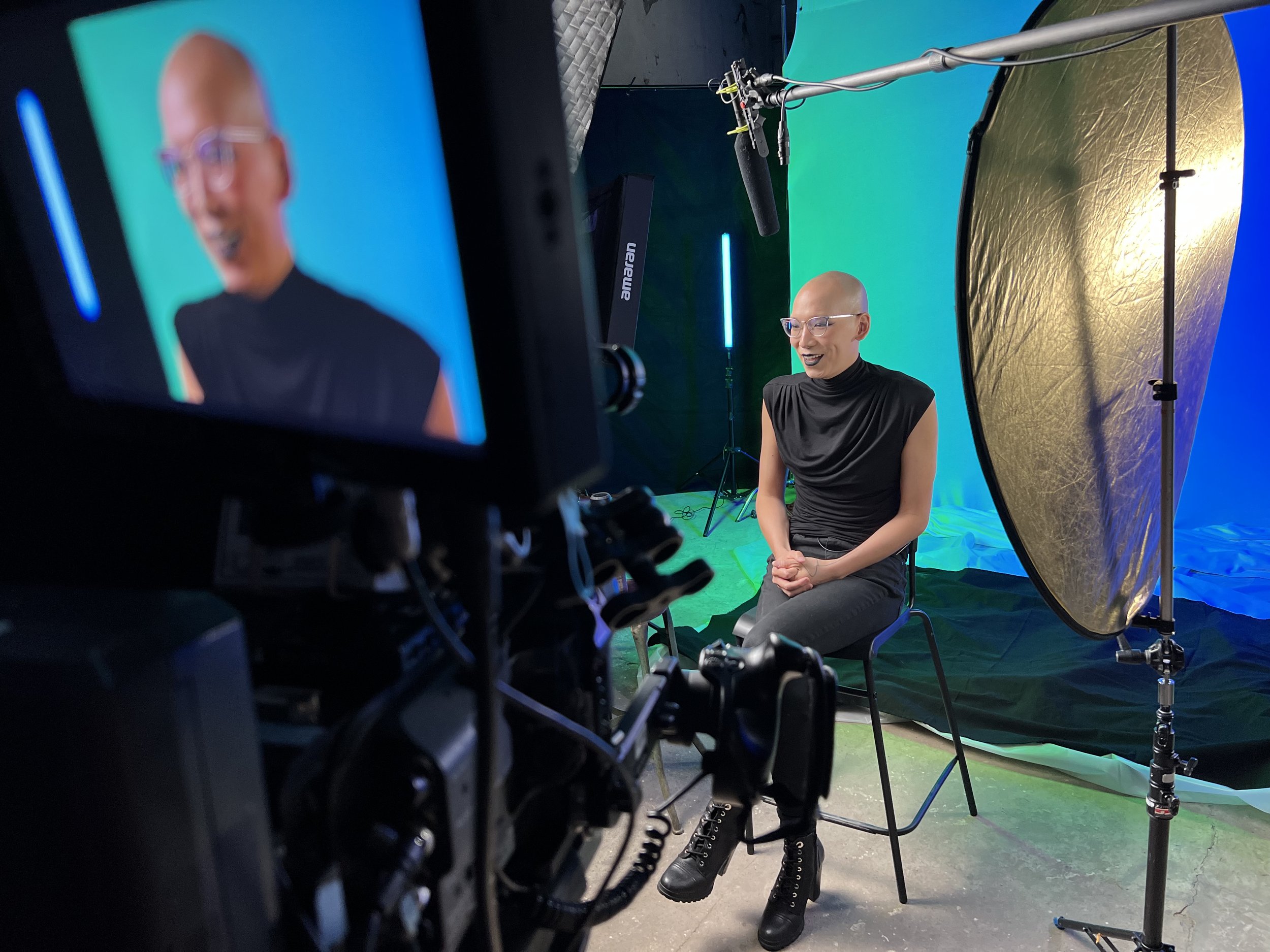 A person with glasses and a shaved head sitting in a photography studio surrounded by green and gold backdrops and studio lighting equipment.