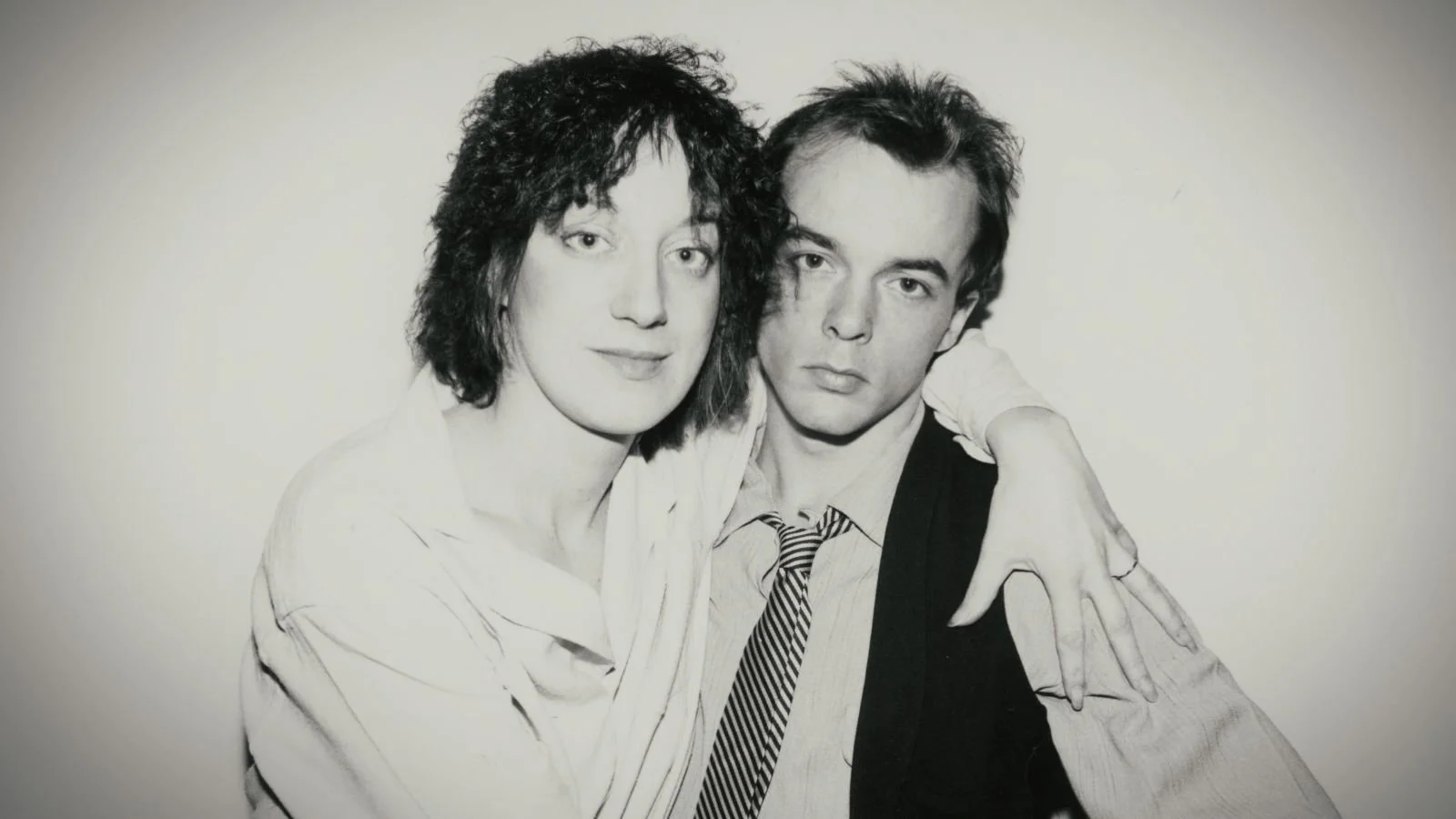Black and white photograph of a woman and a man posing closely, with the woman’s arm around the man's shoulders. The woman has curly hair and a slight smile, while the man has straight hair and a serious expression.