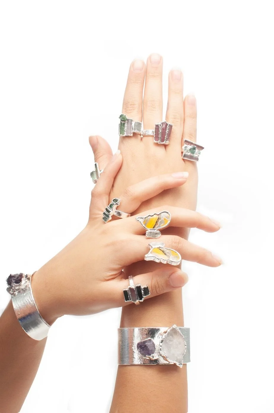 Two hands adorned with multiple silver rings and bracelets, featuring colorful gemstone and abstract designs, against a white background.