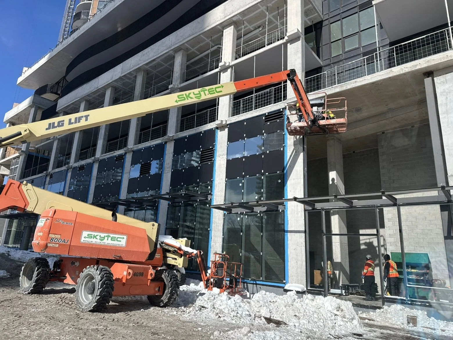 Construction workers working on a high-rise building with glass windows, using a cherry picker lift and scaffolding, on a snowy day.