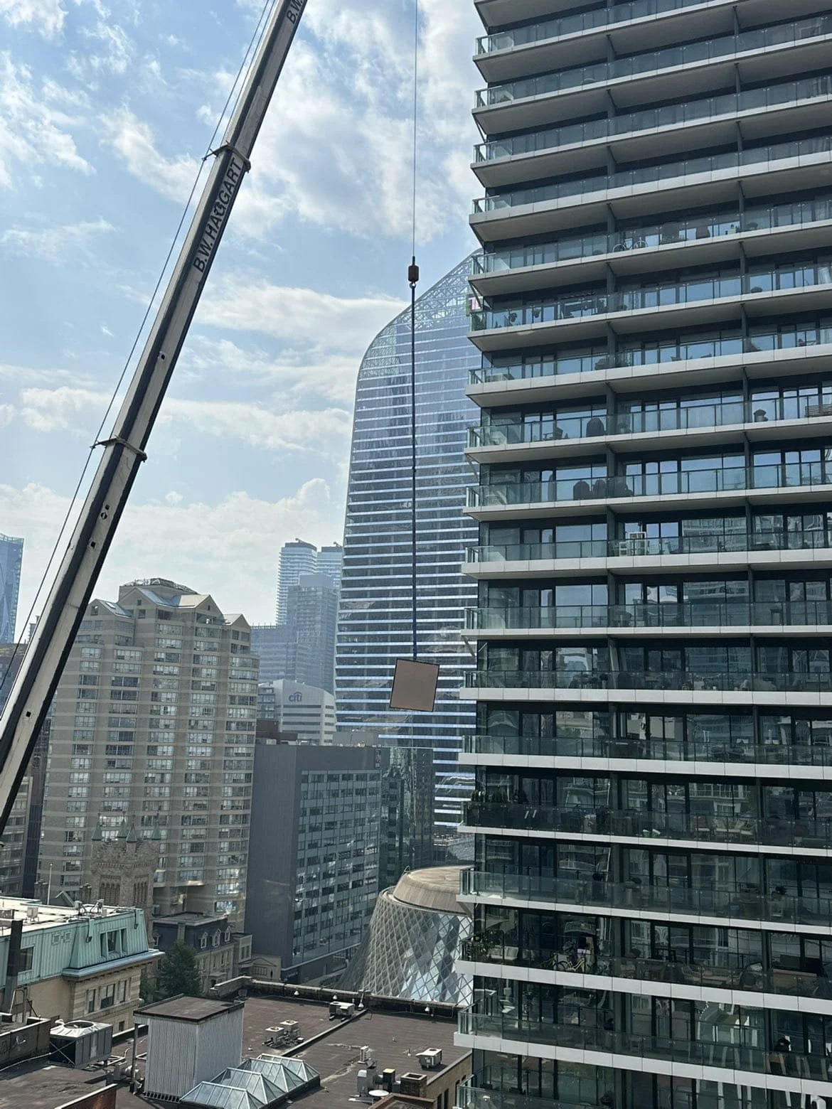 A construction crane lifting a square panel in a city skyline with various tall modern buildings, including a distinctive curved skyscraper, under a partly cloudy sky.