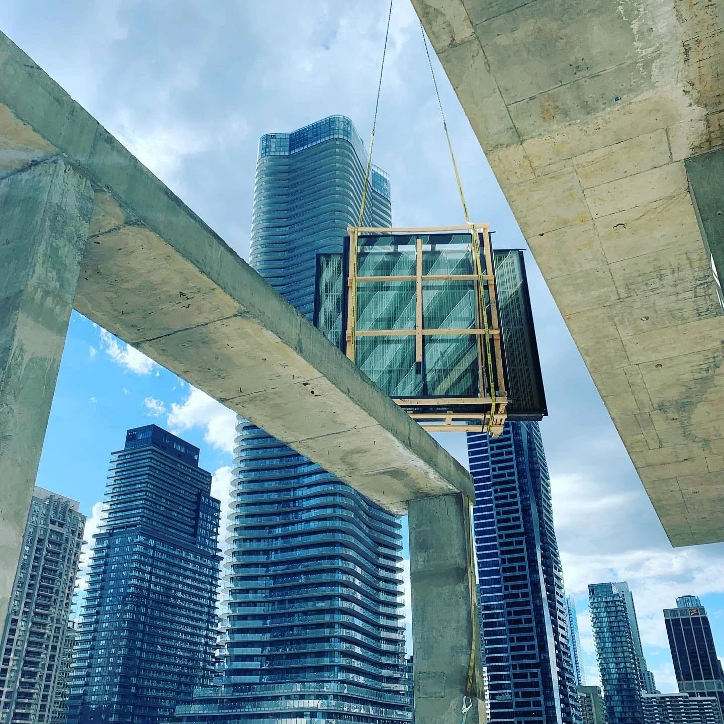 Construction site in a city with multiple high-rise buildings, concrete beams and a glass enclosed platform suspended in the air during daylight.