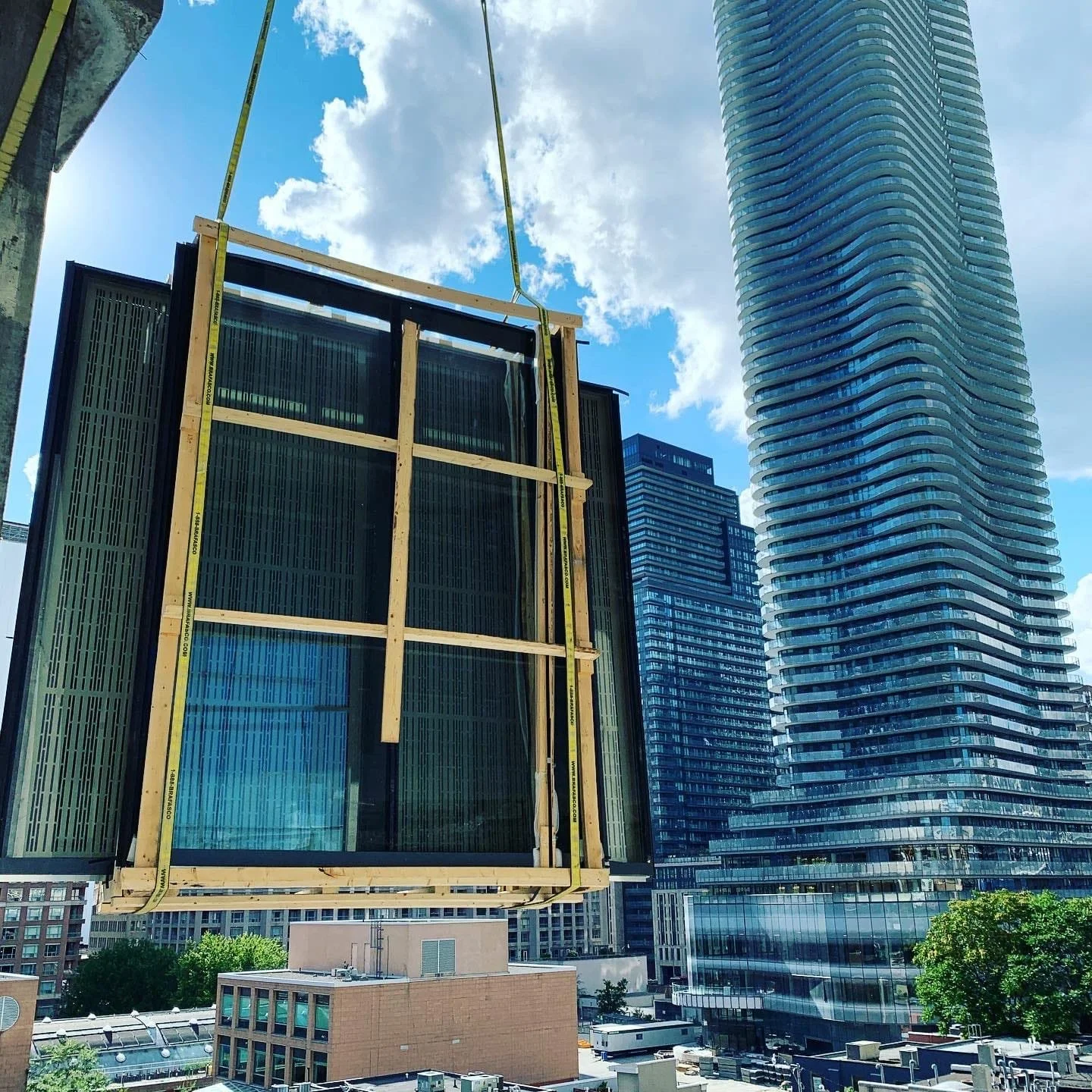 Construction site with a large glass panel being lifted by a crane, surrounded by tall modern skyscrapers under a partly cloudy sky.