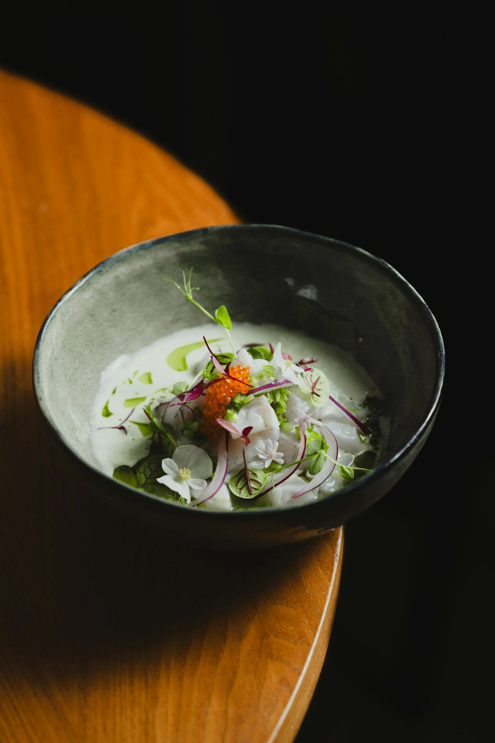 A bowl of creamy soup garnished with microgreens, edible flowers, and fish roe, placed on a wooden table with a dark background.