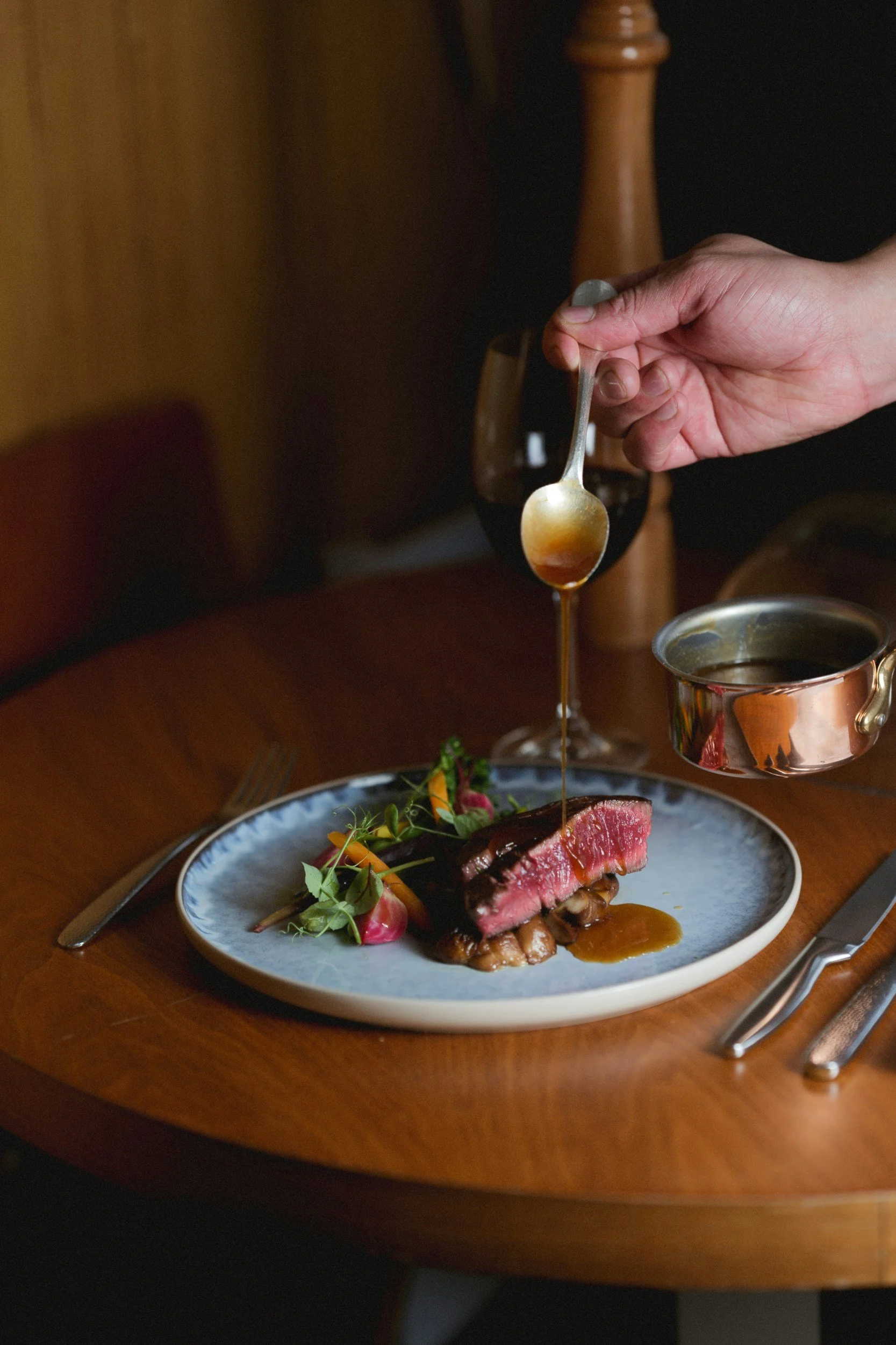 A plated gourmet meal featuring a cooked medium-rare steak with sauce, vegetables, and greens. A hand is pouring sauce from a small pitcher onto the meat.