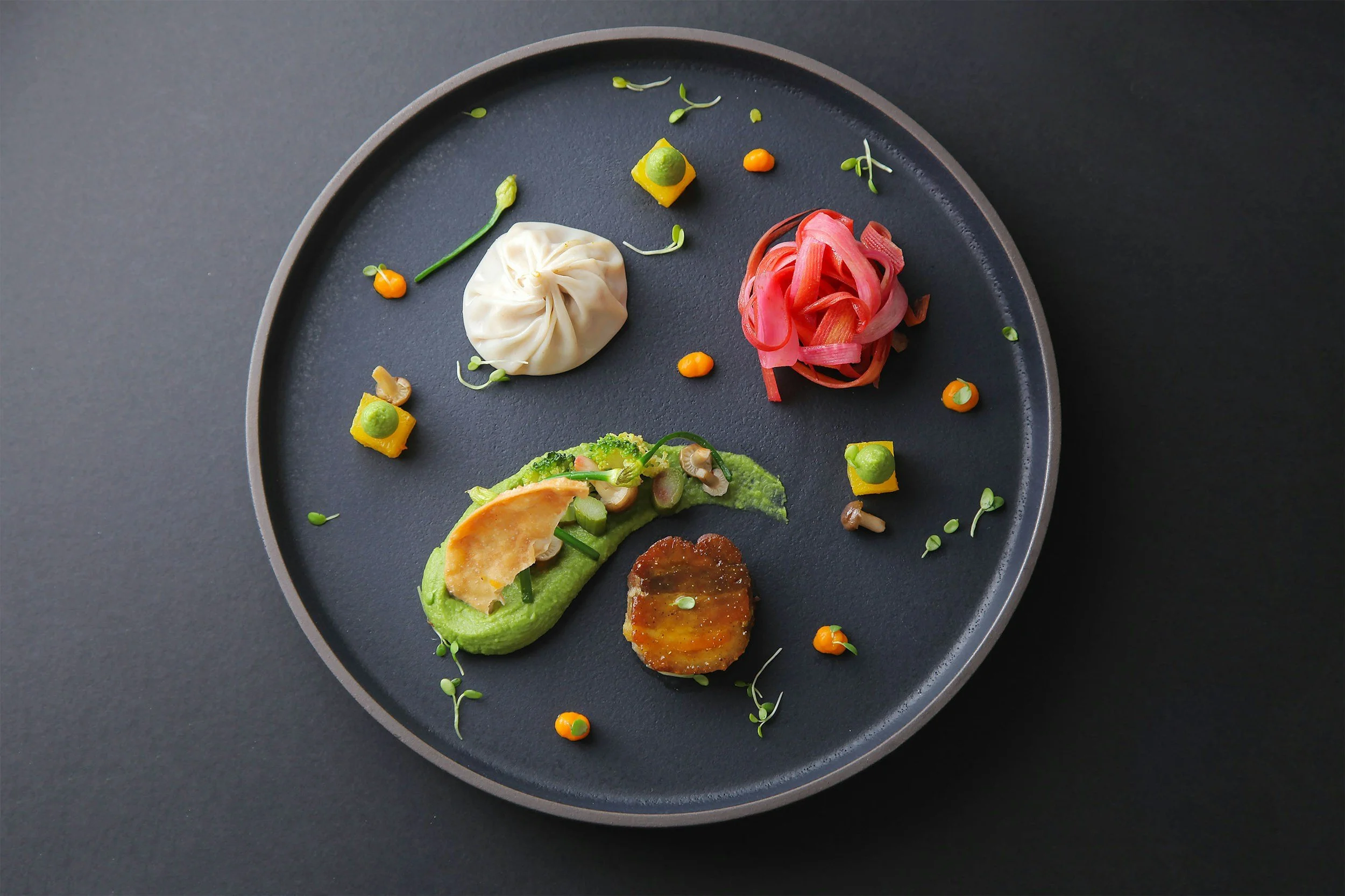 A black round plate with various colorful food items, including a pink pasta nest, a white dumpling, small green and yellow garnishes, and a few small pieces of vegetables, on a dark background.