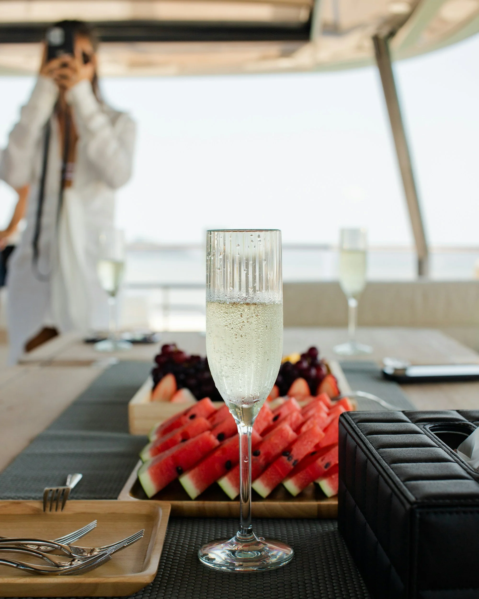 A glass of sparkling water on a dining table with sliced watermelon and a tray of mixed fruits in the background, with a woman taking a photo in the distance.