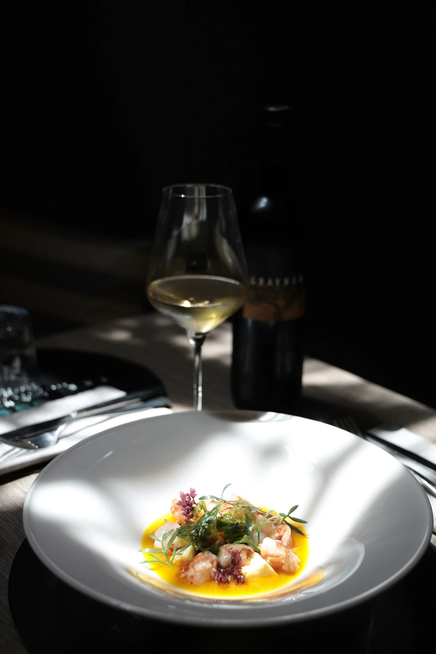 A white bowl with seafood topped with herbs and sauce, set on a table with silverware and a dark background, with a glass of white wine nearby.