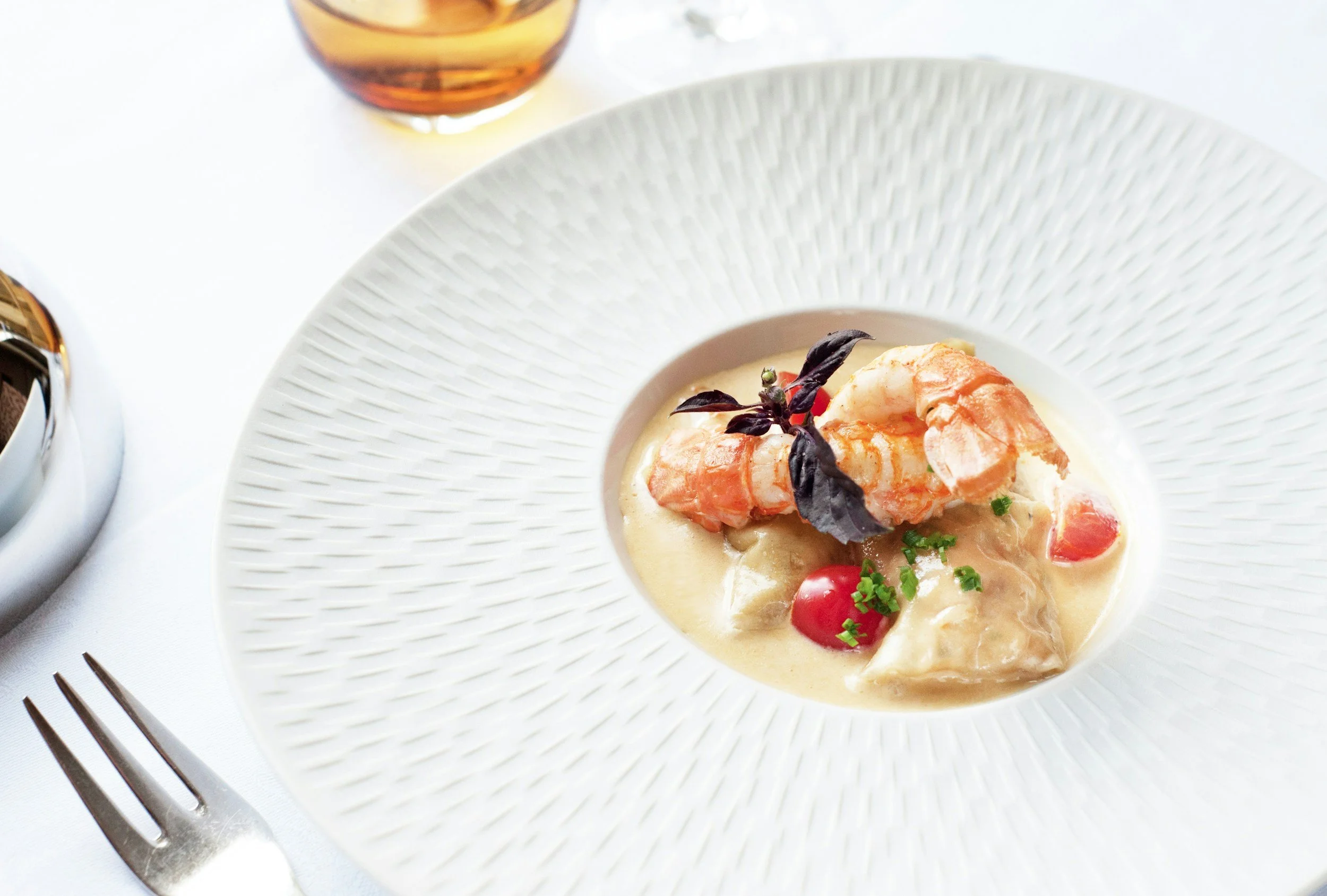 A white plate with a creamy seafood dish topped with shrimp, cherry tomatoes, and purple basil leaves, on a white tablecloth with a fork nearby.