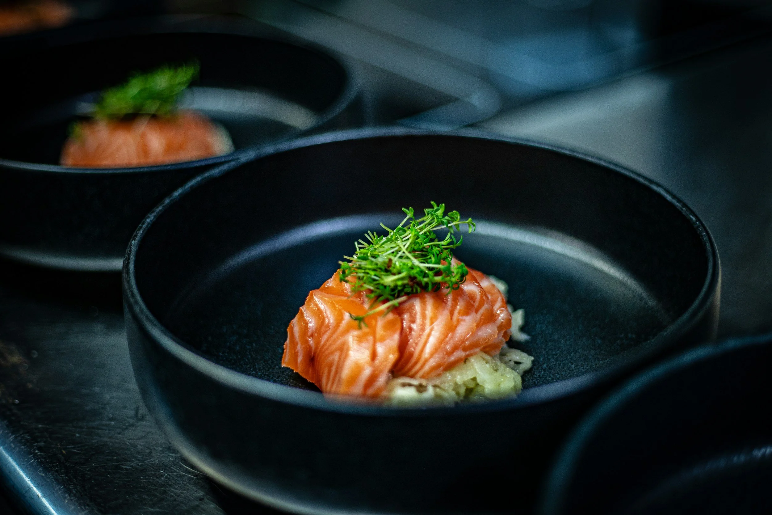 Close-up of a black bowl containing a serving of sashimi-grade salmon topped with fresh microgreens, placed on a dark surface with other similar bowls in the background.