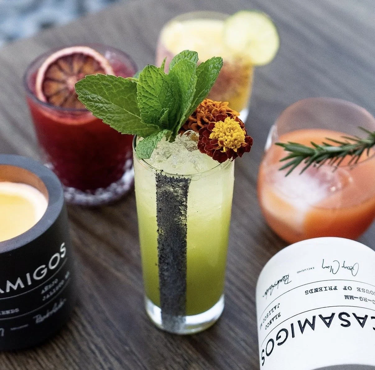 A tall glass of green cocktail garnished with mint leaves, edible flowers, and berries, surrounded by other colorful drinks and a bottle on a wooden surface.