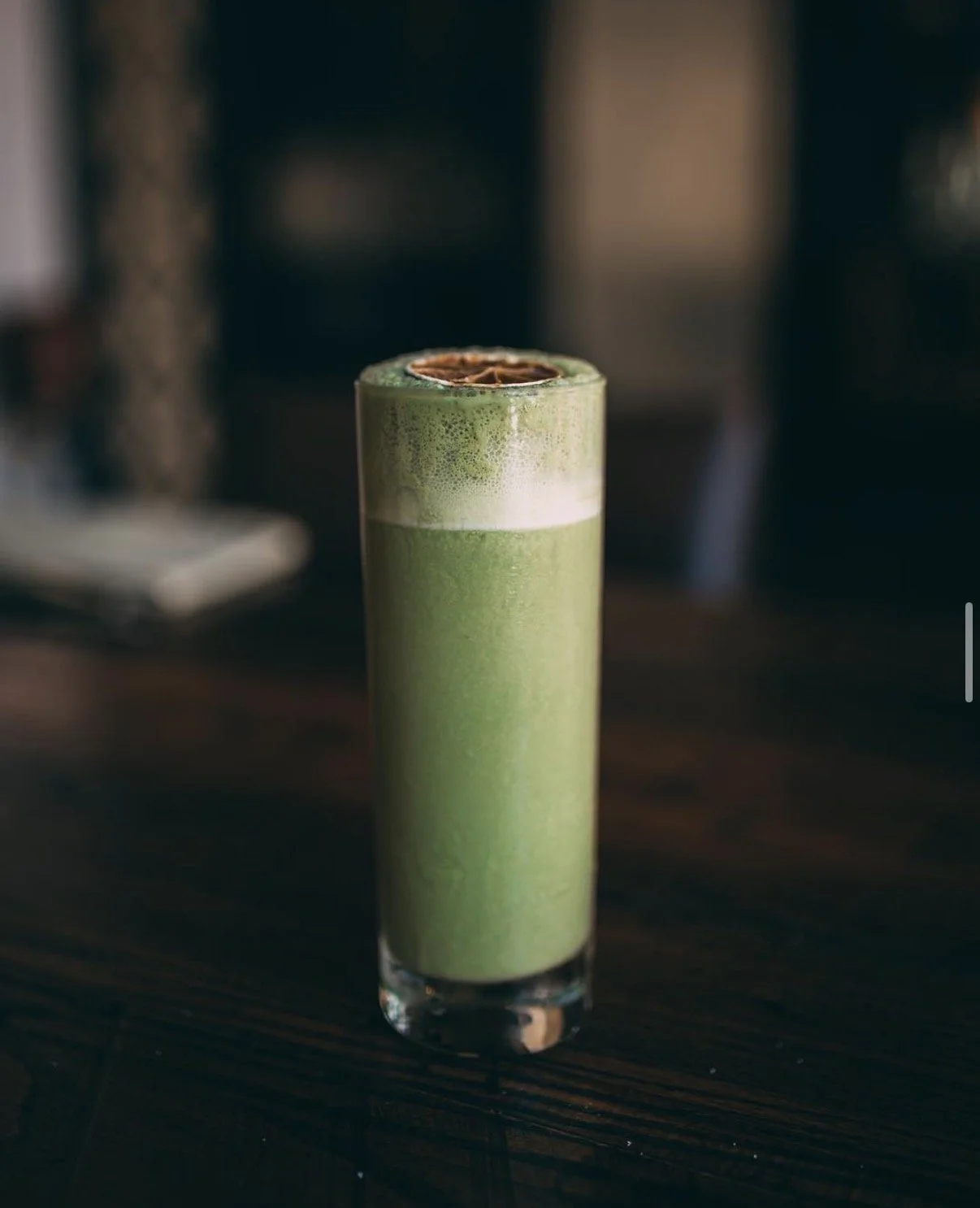 A tall glass of matcha latte with frothy milk and a dusting of matcha powder, topped with a slice of lemon on a dark wooden table.