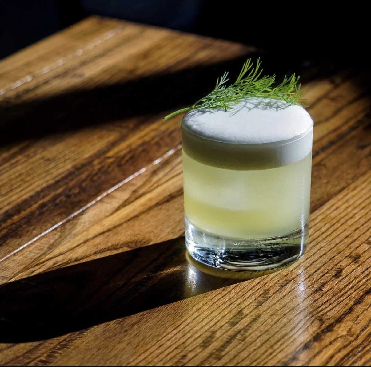A cocktail in a short glass garnished with a sprig of dill on top, placed on a wooden surface with sunlight casting shadows.