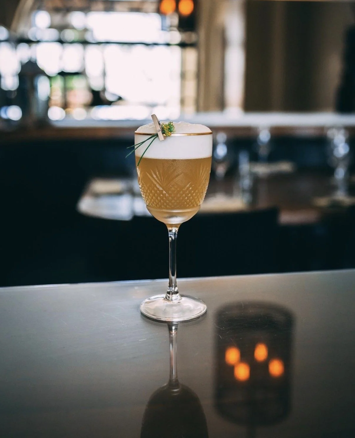 A cocktail glass filled with a golden drink topped with foam, garnished with a small decorative flower and greenery, on a reflective dark surface in a dimly lit bar or restaurant.