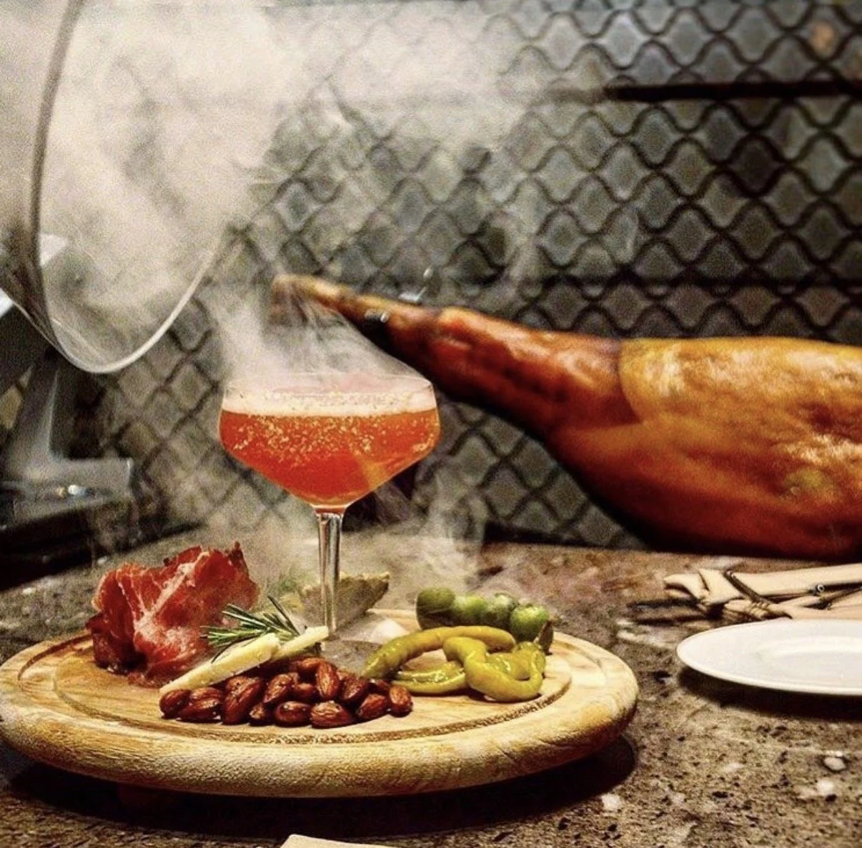 A smoked cocktail with smoke coming out, a charcuterie board with almonds, prosciutto, garlic, rosemary, and peppers, a large smoked ham, and a glass of beer on a countertop.
