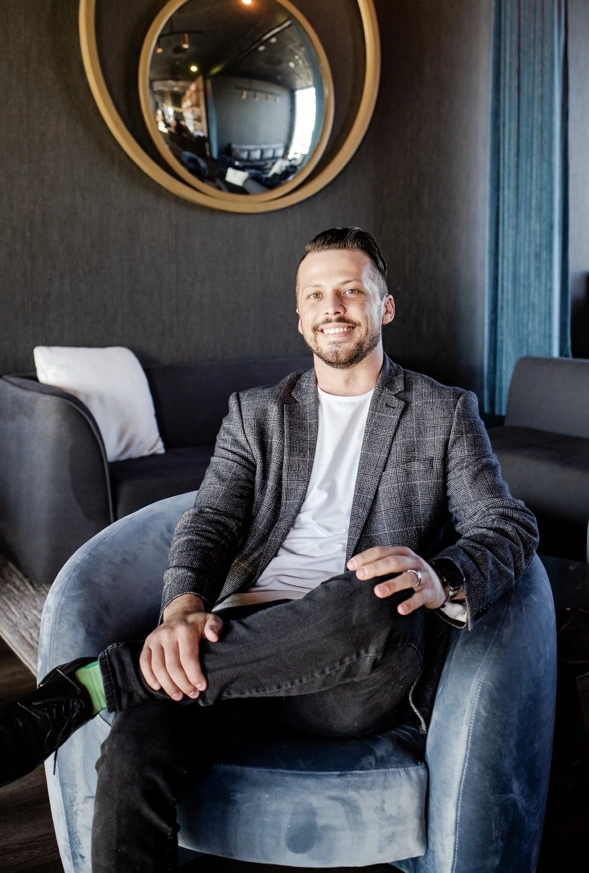 A young man with a beard and short, slicked-back hair sitting in a blue velvet armchair, smiling at the camera in a modern room with dark walls, a large mirror, and a dark sofa in the background.